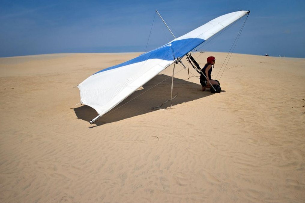 A student pilot kneeling beside a blue and white hang glider on the open sand at Jockey's Ridge