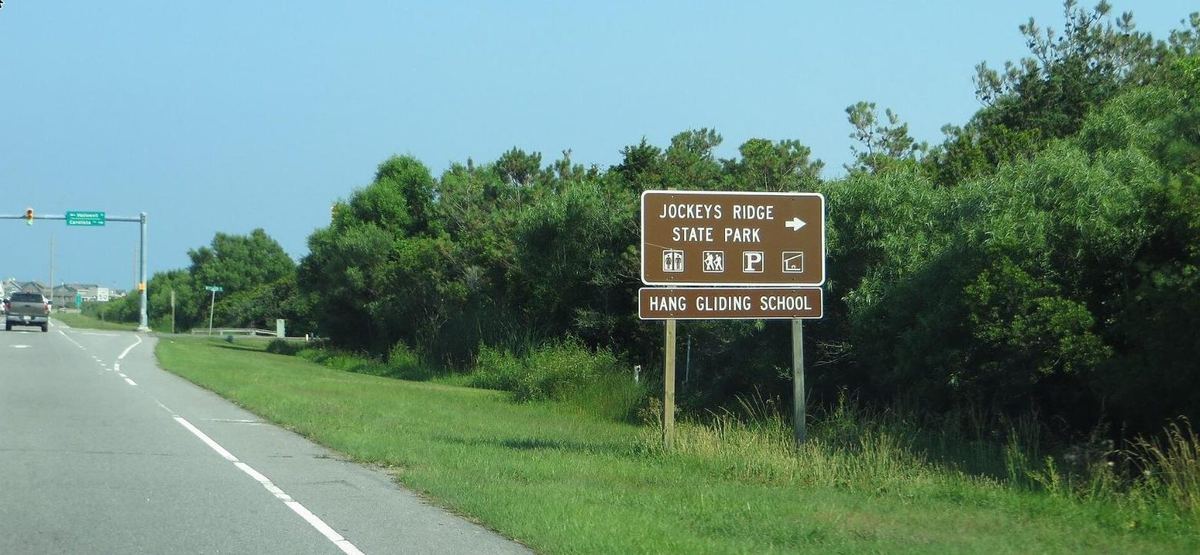 A brown highway sign reading Jockeys Ridge State Park with a second panel reading Hang Gliding School