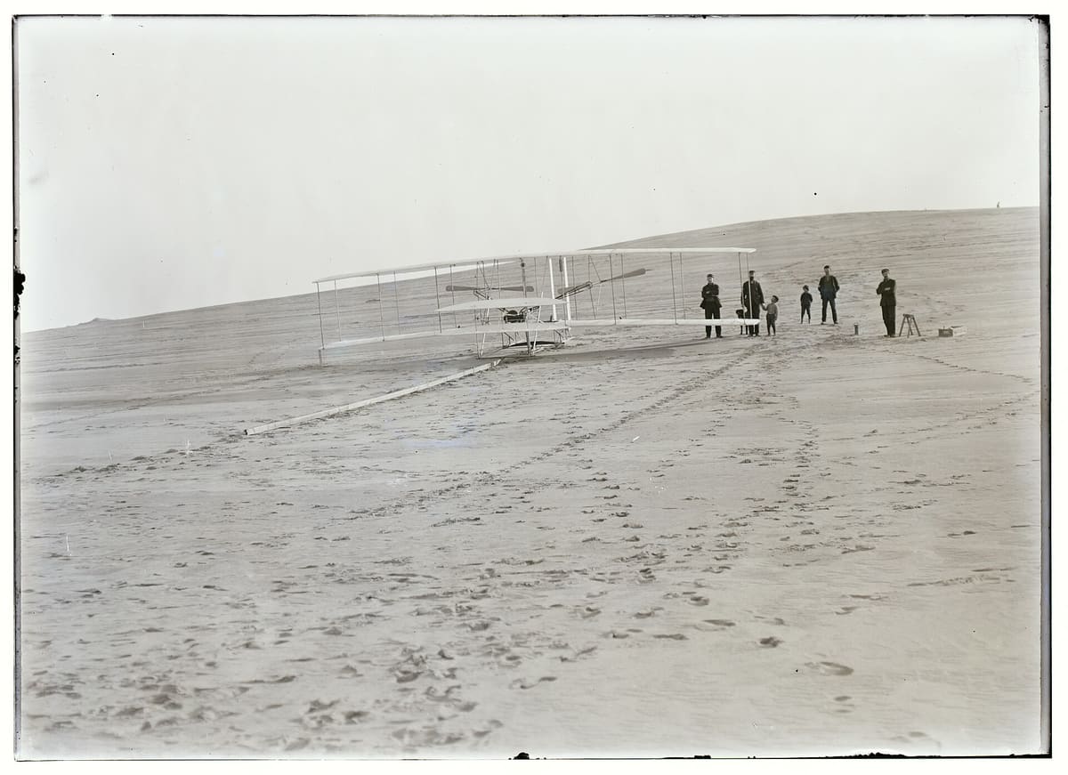 The 1903 Flyer sitting on its wooden launch rail on open sand, with several men standing beside it